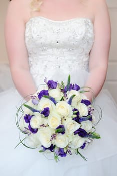 Bride holding bouquet of white and purple flowers on wedding day showing dress detail closeup