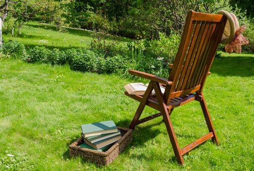 wooden relax chair, books in basket and hat in garden yard in summer day.