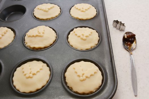 Mince pies ready to be baked, with teaspoon of mincemeat and a holly leaf cutter on the worktop