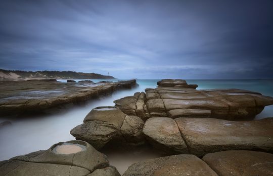 Weathered rocks, ocean channel and rock craters at Soldiers Beach Australia under a stormy dark sky.  Norah Head lighthouse in distance.
