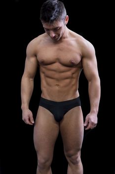 Muscular young man semi naked in bathing suit looking down, isolated on black