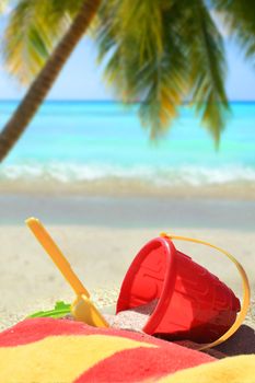 Beach toys and towel in red and yellow in  sand on a tropical Caribbean beach with palm tree and blue ocean in the background      