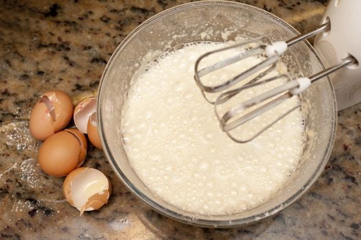 View from above of broken eggshells and a frothy batter mix in a metal mixing bowl on an electric mixer with whisk attachments for baking