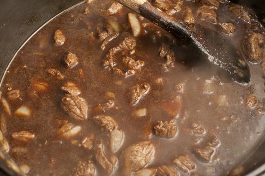 Pot of rich brown beef stew with multiple cubes of meat in a seasoned gravy simmering on a stove while being stirred with a wooden spoon