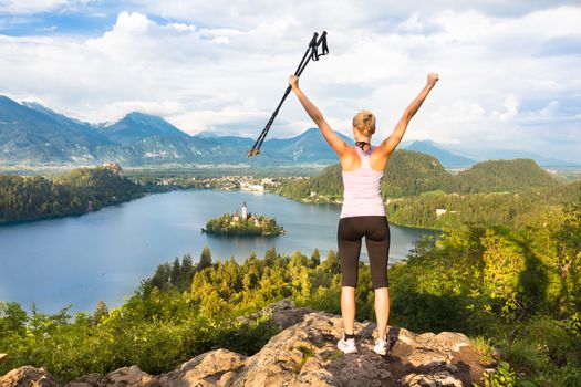 Made it! Young spoty active lady with hiking sticks and hands rised admiring beautiful nature around Bled Lake in Julian Alps, Slovenia. 