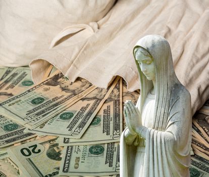 Image of statue of lady praying with US currency as background as a concept for the linkage of religion with money and wealth
