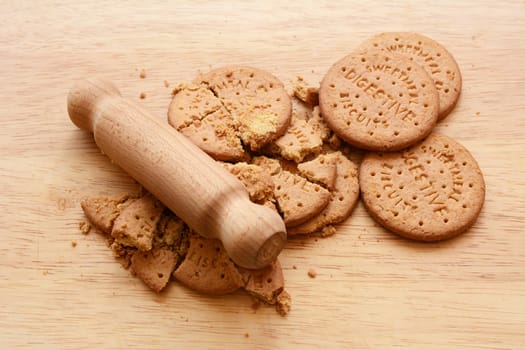 Whole and crushed digestive biscuits on a chopping board with a small wooden rolling pin