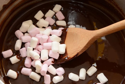 Stirring pink and white mini marshmallows into melted chocolate with a wooden spoon to make rocky road