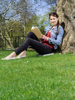 Photo of a beautiful young woman reading a book sitting against a tree in early spring.