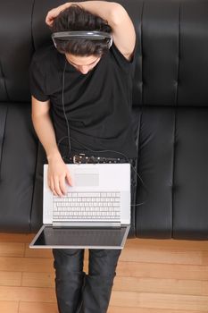 A young hispanic man with a laptop on the Sofa listening Music with headphones.