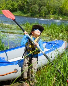 Portrait of happy young boy holding paddle near a kayak on the river, enjoying a lovely summer day