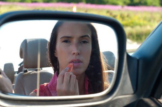 Young caucasian woman applying lipstick looking at reflection in car mirror
