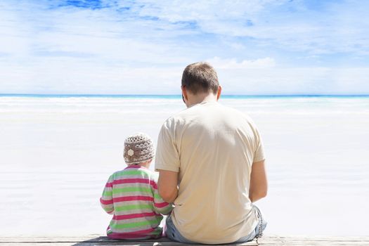 Father and daughter sitting on empty white sand beach with turquoise ocean on sunny day. Relationship, family, lifestyle, fatherhood, togetherness.