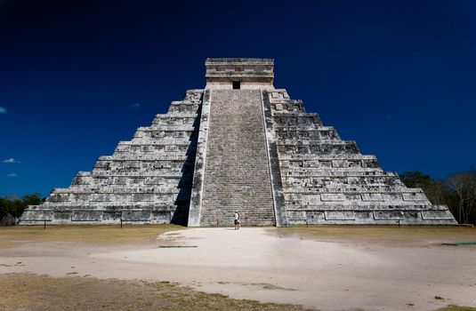 Ziggurat (pyramid) at Chichen Itza with two people staring at it in front of the stairway. Dark blue sky