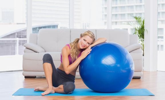 Toned blonde sitting beside exercise ball smiling at camera at home in the living room