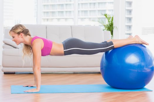 Slim blonde in plank position using exercise ball at home in the living room