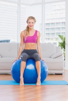 Toned blonde sitting on exercise ball smiling at camera at home in the living room
