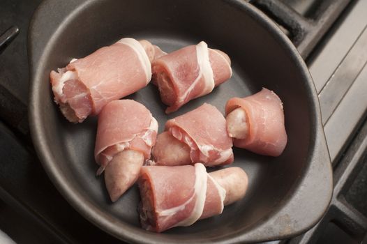 Cooking pigs in blankets, or rashers of bacon wrapped around beef and pork sausages, with a batch of uncooked piggies in a cast iron casserole dish waiting to be cooked in the oven or on a hot griddle