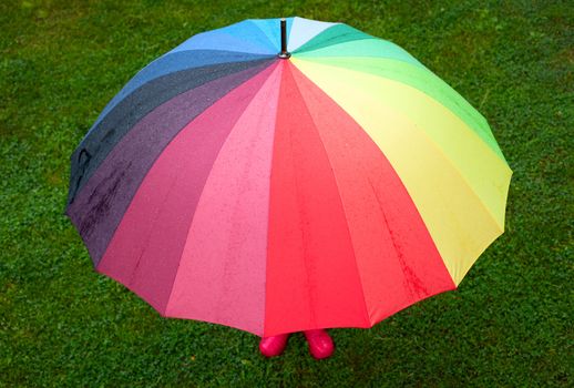 Little girl wearing red rubber boots hiding behind colorful umbrella in the rain