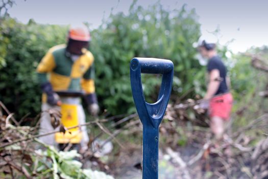 Close up of a handle of a garden fork with two male figures in the background in soft focus maintaining a garden in protective head gear. Set on a landscape format.