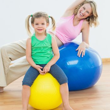 Smiling pregnant woman exercising on exercise ball with young daughter in a fitness studio