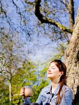 Photo of a beautiful young woman drinking coffee or tea while sitting outdoors enjoying the warmth of the sun on her face.