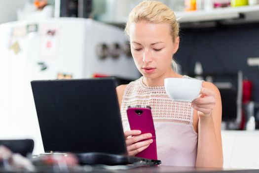 Business woman working from her home. Multitasking. Having morning coffee while texting on the smarth phone and working on leptop computer.