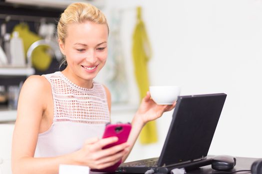Business woman working from her home. Multitasking. Having morning coffee while texting on the smarth phone and working on leptop computer.