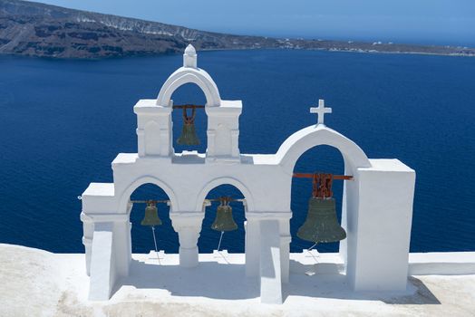 View On Calm Sea Surface Through Traditional Greek White Church Arch With Cross And Bells In Village Oia of Cyclades Island Santorini, Greece