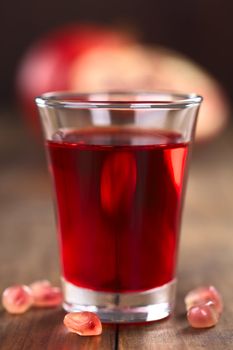 Grenadine syrup in shot glass with pomegranate seeds on the side and pomegranate in the back (Selective Focus, Focus on the front rim of the glass)