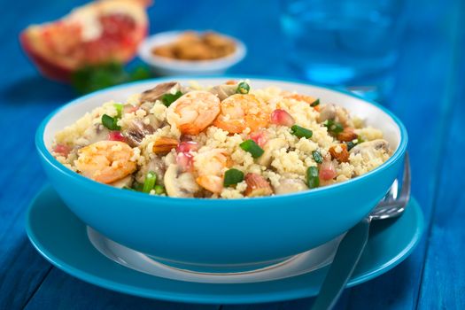 Couscous dish with shrimps, mushroom, almond, pomegranate seeds and green onion served in blue bowl with fork on the side (Selective Focus, Focus on the tails of the shrimps on the top of the meal) 
