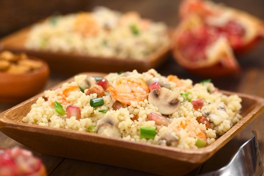 Couscous dish with shrimps, mushroom, almond, pomegranate seeds and green onion served on wooden plate (Selective Focus, Focus on the tail of the shrimp on the top of the meal and on the mushroom slice next to it) 