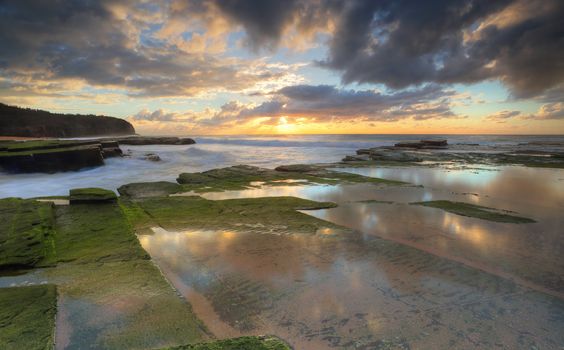 The riising sun hides behind some clouds.  The Turrimetta coastline and reflectios during winter Sydney Australia