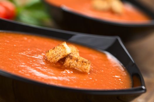 Fresh homemade tomato soup with wholegrain croutons on top served in black bowl on dark wood (Selective Focus, Focus on the croutons in the front) 