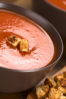 Fresh homemade tomato soup with wholegrain croutons on top in brown bowl on dark wood (Selective Focus, Focus on the front of the croutons on the soup) 
