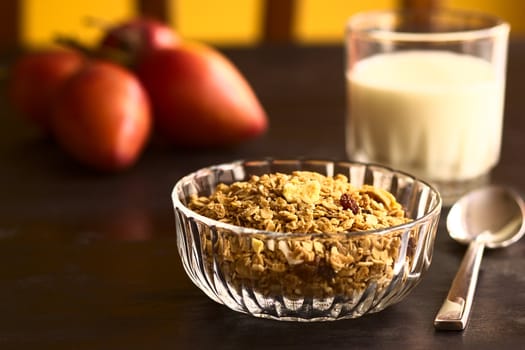 Breakfast cereal out of oat bran flakes, sesame, honey, almonds and dried fruits (coconut, apple, banana, raisins) in glass bowl with a glass of milk and tamarillo in the back (Selective Focus, Focus on the dried fruit pieces on the top of the cereal)