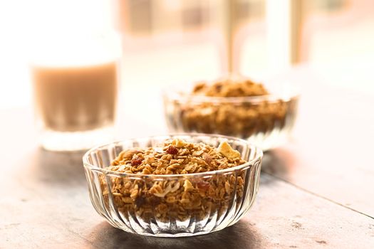 Breakfast cereal out of oat bran flakes, sesame, honey, almonds and dried fruits (coconut, apple, banana, raisins) in glass bowl with a glass of milk in the back (Selective Focus, Focus on the raisins on the top of the cereal)