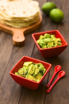 Two red bowls of fresh avocado salad prepared with lime juice, pepper, salt and garnished with fresh coriander leaves, homemade tortillas in the back (Selective Focus, Focus in the middle of the salad)  