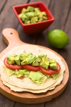 Fresh homemade tortilla with lettuce, tomato and avocado on wooden board with avocado salad and lime in the back (Selective Focus, Focus on the front of the avocado pieces on top)