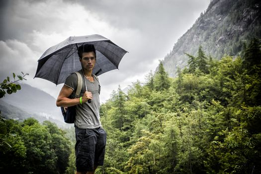 Smiling handsome young man holding an umbrella on a stormy day as he takes a walk along a trail in the mountains, looking at camera
