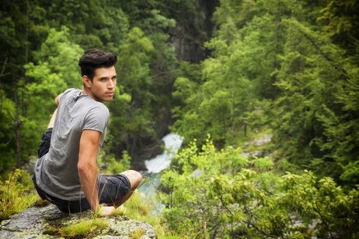 Handsome muscular young man sitting in lush green mountain scenery looking at camera