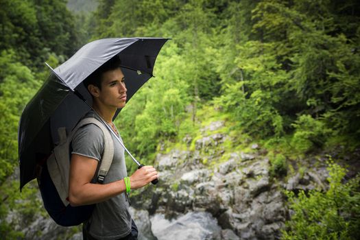 Smiling handsome young man holding an umbrella on a stormy day as he takes a walk along a trail in the mountains, looking far away