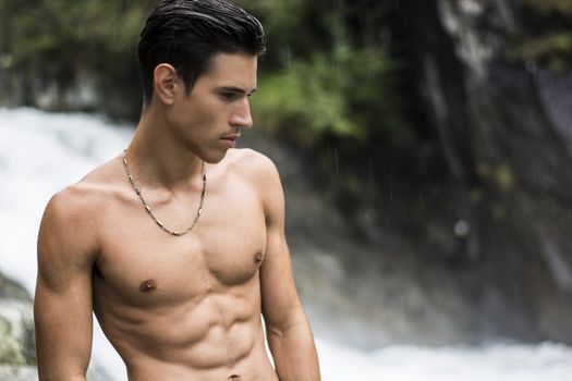 Handsome shirtless young man near mountain waterfall, with ripped fit body