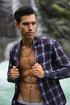 Handsome young man near mountain waterfall with open shirt, ripped athletic body