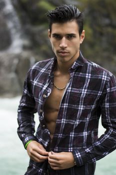 Handsome young man near mountain waterfall with open shirt, ripped athletic body