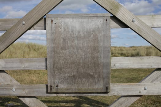 A blank wooden sign attached to a wooden framework with grass and a blue cloudy sky in the background.