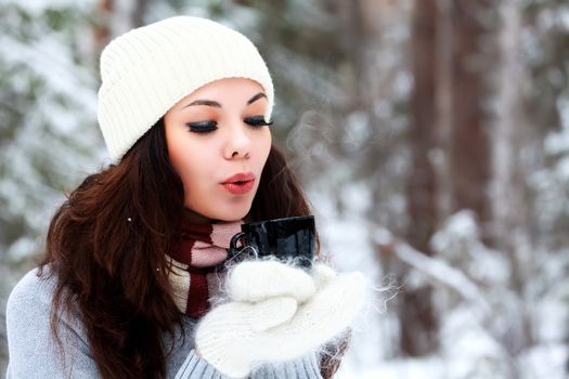 Beautiful young woman in knitted hat and sweater blows on hot tea or coffee
