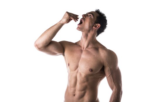 Strong muscular man taking diet supplements or pharmaceutical medication standing shirtless with his mouth open and head tilted back as he drops a pill from his hand or places something in his mouth