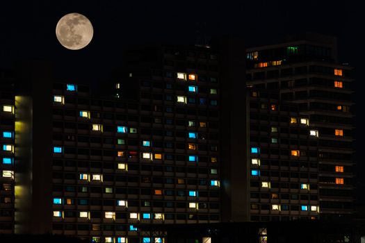 Full moon in night sky over colorful illuminated windows of residential high rise apartments house