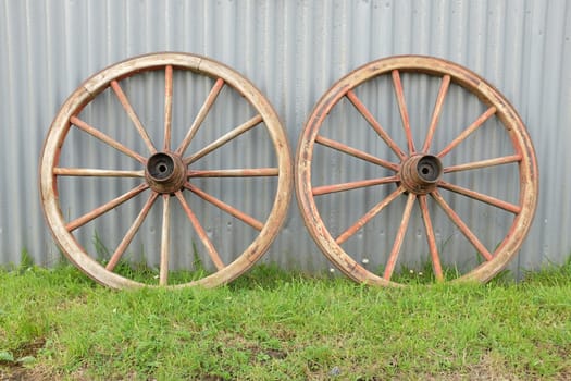 Green grass leads towards a pair of wooden made antique cart wheels resting against a metal wall.
